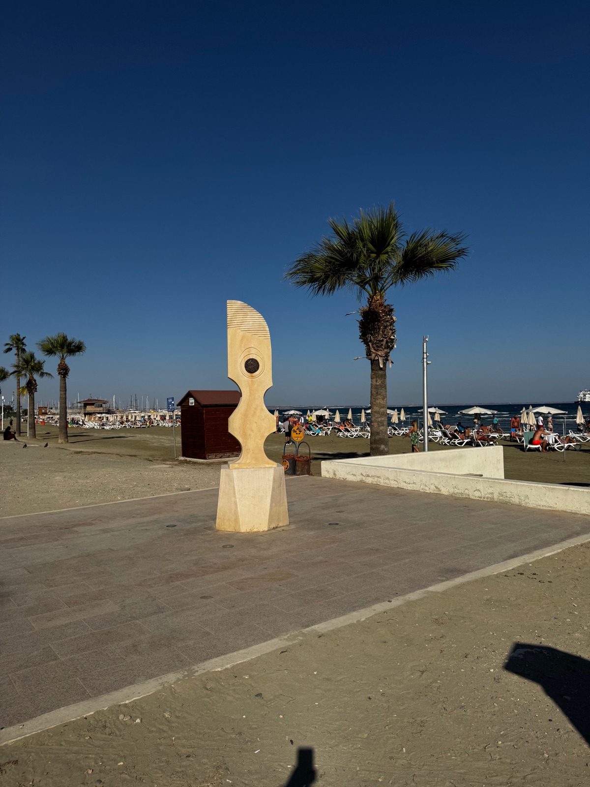 Ένα χλωμό αφηρημένο πέτρινο γλυπτό με στρογγυλό μπρούντζινο δίσκο να στέκεται στον περίπατο της Λάρνακας, φοίνικες και ομπρέλες παραλίας πίσω