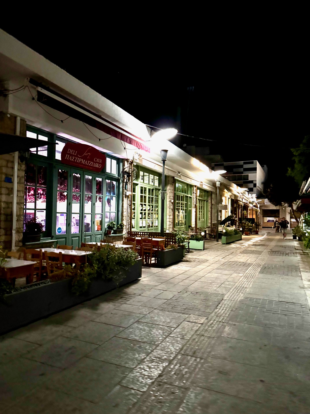 Ένας άδειος πεζόδρομος στην παλιά πόλη της Λεμεσού τη νύχτα, τραπέζια ταβέρνας και παράθυρα με πράσινα κουφώματα κάτω από μια κόκκινη πινακίδα ντελικατέσεν στα ελληνικά