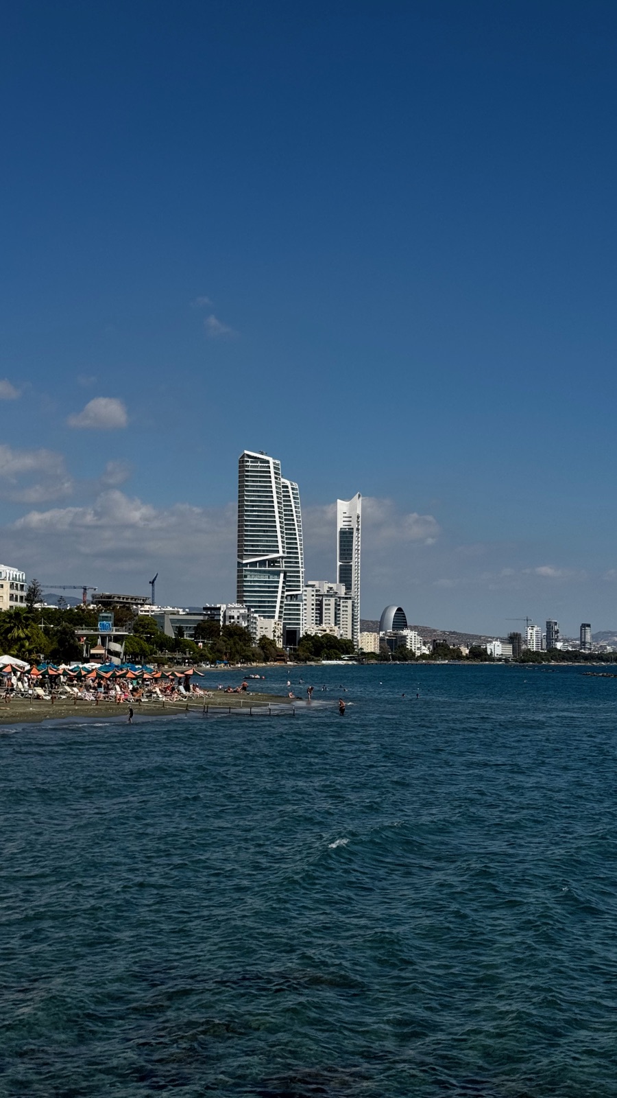 Μια γεμάτη παραλία της Λεμεσού τον Οκτώβριο με πορτοκαλί ομπρέλες, δύο καμπύλοι πύργοι και ένας λευκός ουρανοξύστης να υψώνονται πίσω