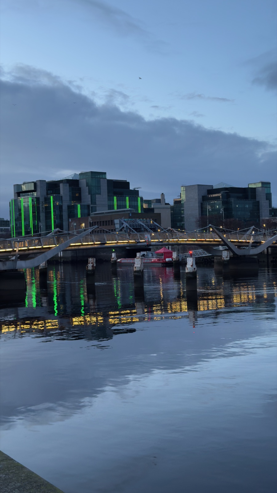 Κτίρια γραφείων στα docklands του Δουβλίνου φωτισμένα πράσινα το σούρουπο δίπλα σε γέφυρα, να καθρεφτίζονται στο ήρεμο ποτάμι
