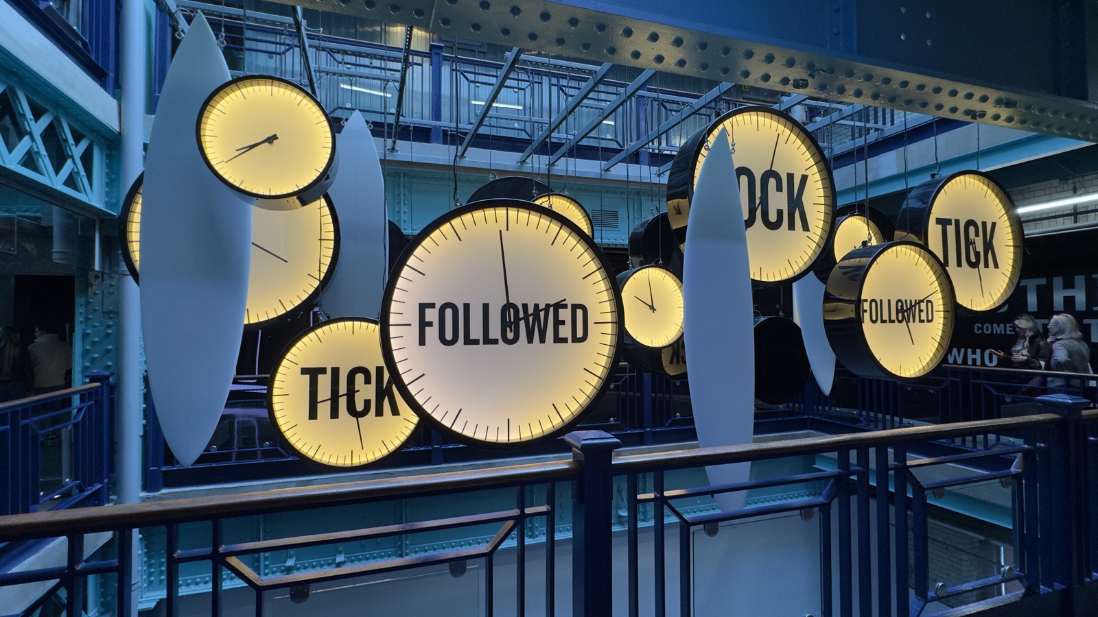 Εγκατάσταση με μεγάλα φωτισμένα ρολόγια και λευκά σχήματα σαν σανίδες σερφ κρεμασμένα πάνω από μπλε μεταλλικό κλιμακοστάσιο, που συνθέτουν τις λέξεις tick, tock και followed
