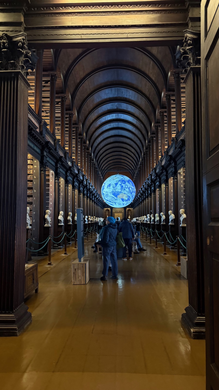 Το Long Room της Old Library του Trinity College, μια μακριά καμάρα από σκούρα δρυ πάνω από δύο σειρές ραφιών και μαρμάρινες προτομές, με μια μεγάλη φωτισμένη υδρόγειο κρεμασμένη στο βάθος
