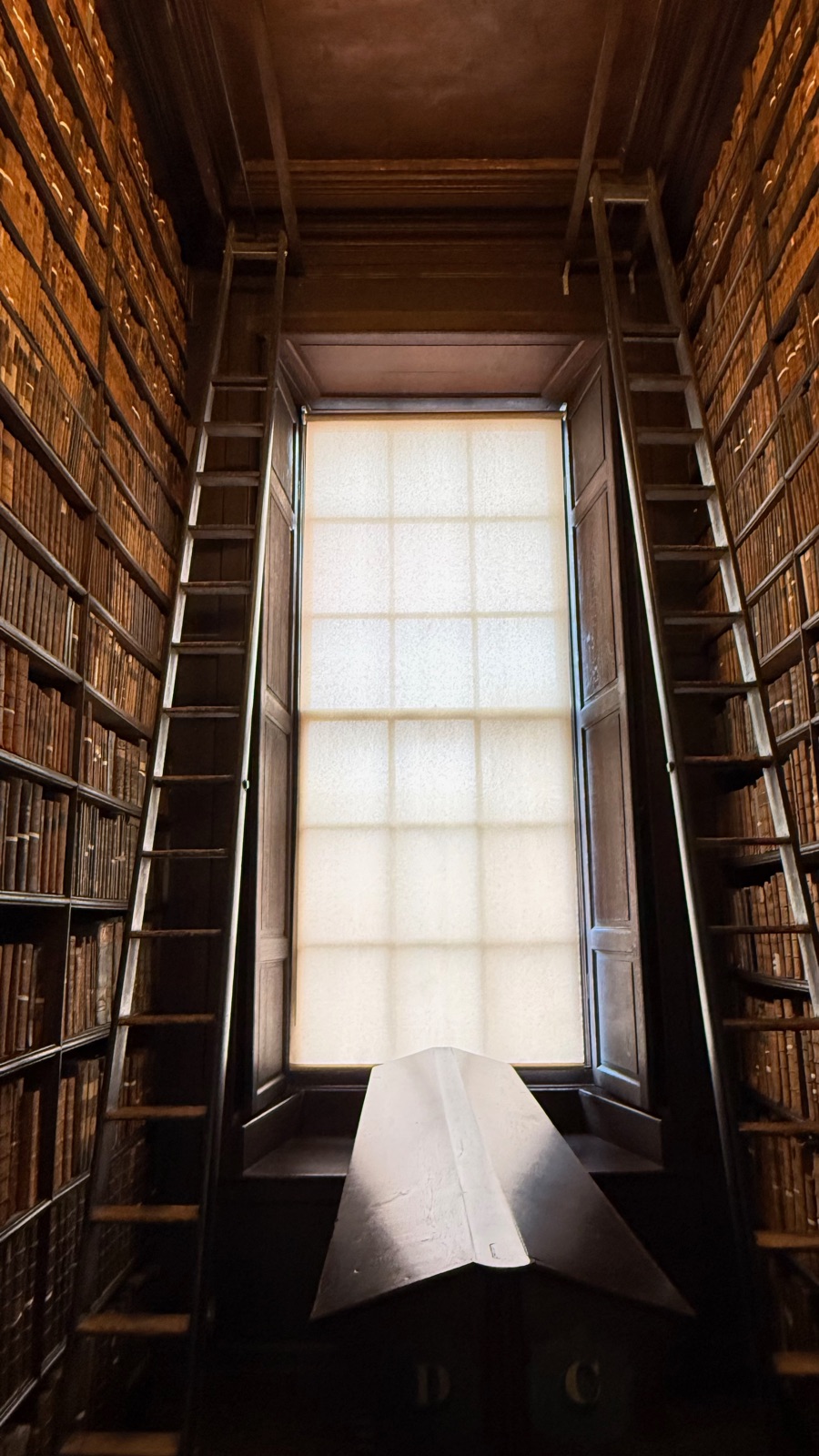 Φατνώμα ραφιών στο Long Room φωτισμένο από ψηλό παράθυρο με φως από πίσω, ένα ανοιχτό βιβλίο σε αναλόγιο ανάμεσα στα ράφια