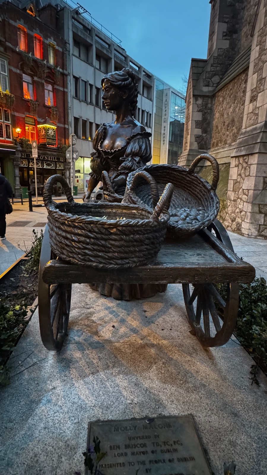 Το χάλκινο άγαλμα της Molly Malone το σούρουπο, μια γυναίκα πίσω από ένα καρότσι με ψάθινα καλάθια, φωτισμένα παράθυρα παμπ και πέτρινος τοίχος πίσω της