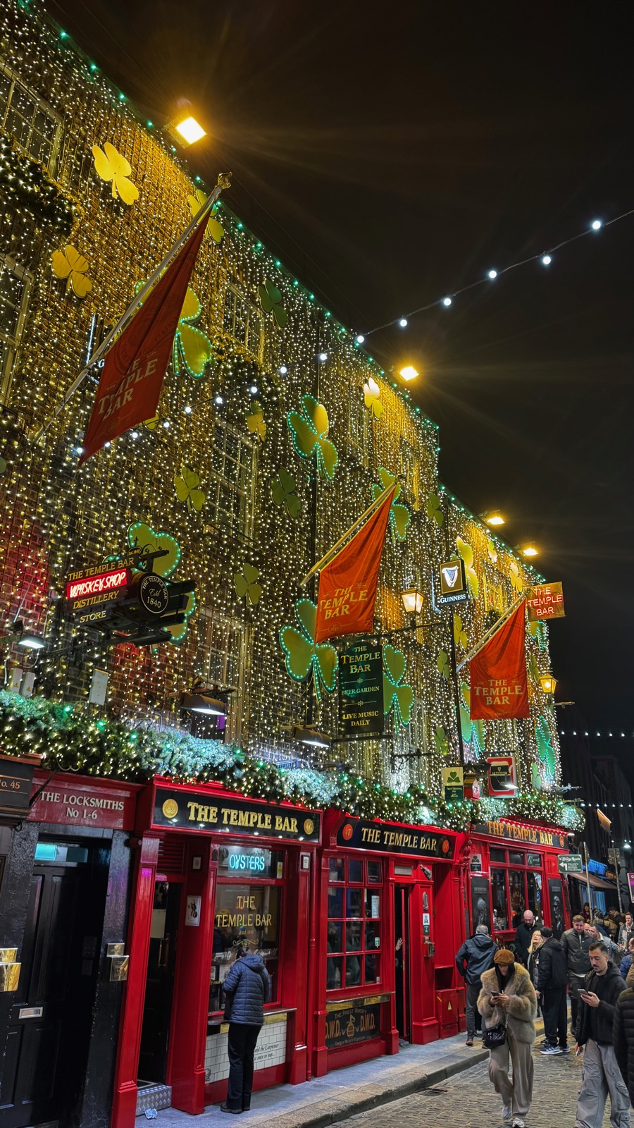 Η παμπ Temple Bar τη νύχτα, όλη της η πρόσοψη καλυμμένη με μικροσκοπικά πράσινα και χρυσά λαμπάκια και πορτοκαλί σημαίες, η κόκκινη βιτρίνα φωτισμένη από κάτω