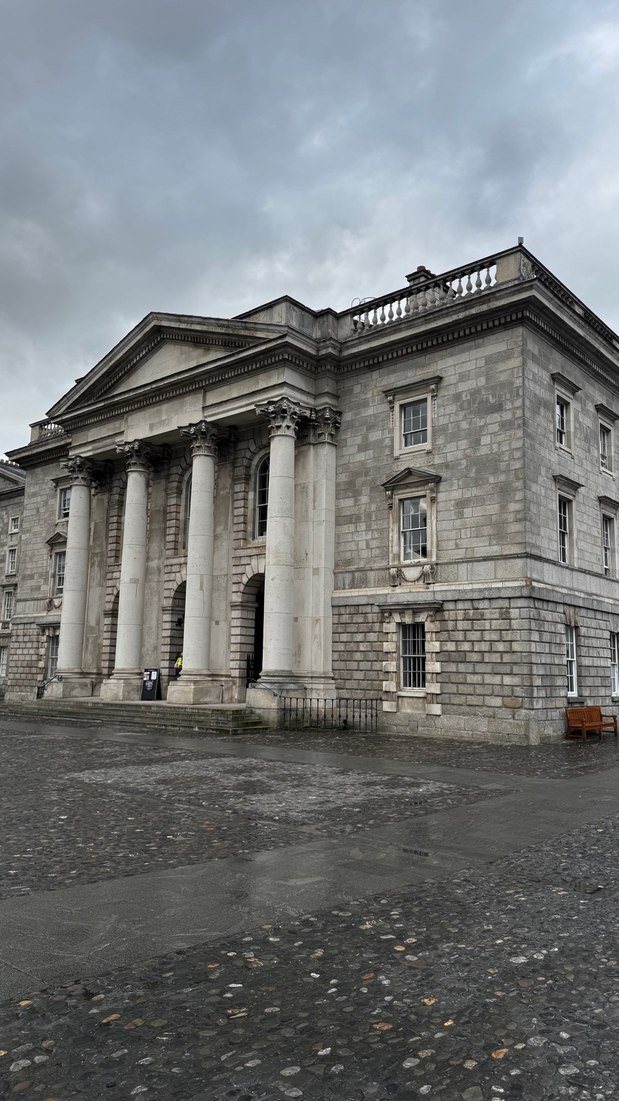 Το γκρίζο πέτρινο πρόπυλο του Exam Hall στο Trinity College, ψηλές κολόνες κάτω από αέτωμα, βρεγμένα βότσαλα μπροστά