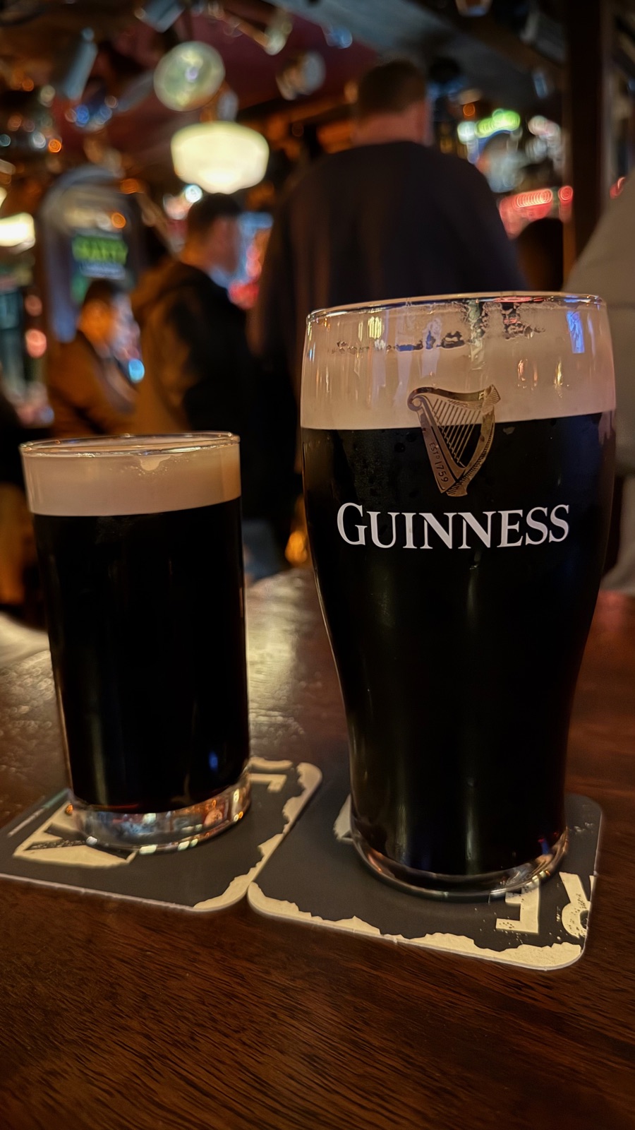 Δύο πίντες Guinness πάνω σε σουβέρ σε σκούρο ξύλινο τραπέζι παμπ, οι κρεμώδεις αφροί καθισμένοι επίπεδα, τα φώτα της παμπ θολά πίσω