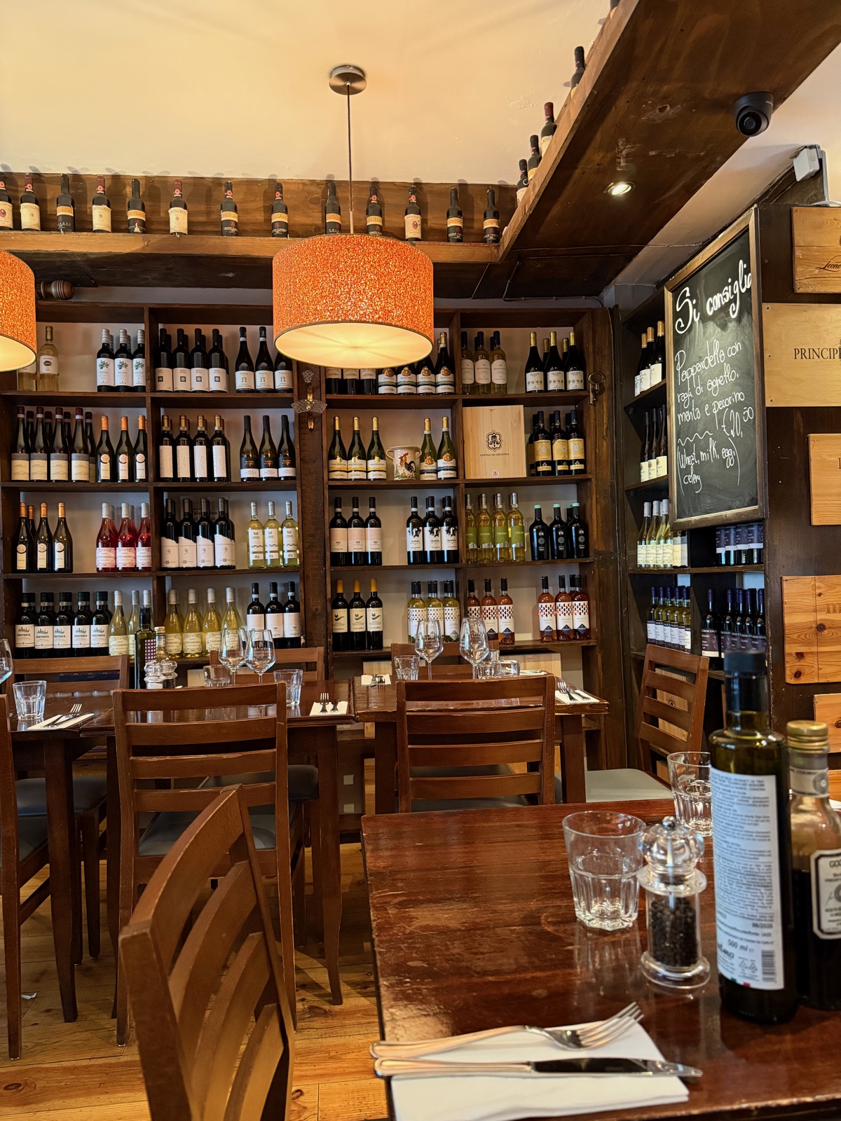 Το εσωτερικό ενός ήσυχου wine bar, ξύλινα τραπέζια στρωμένα για μεσημεριανό κάτω από πορτοκαλί κρεμαστά φωτιστικά, ράφια με μπουκάλια σε όλον τον πίσω τοίχο
