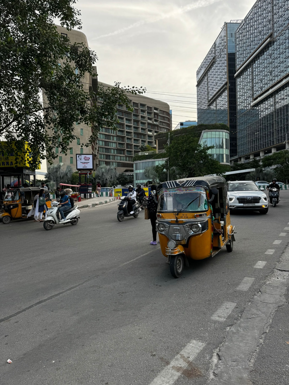 Δρόμος την ημέρα με ένα κίτρινο auto-rickshaw, σκούτερ και μια πινακίδα της Τροχαίας του Cyberabad μπροστά από γυάλινα γραφεία