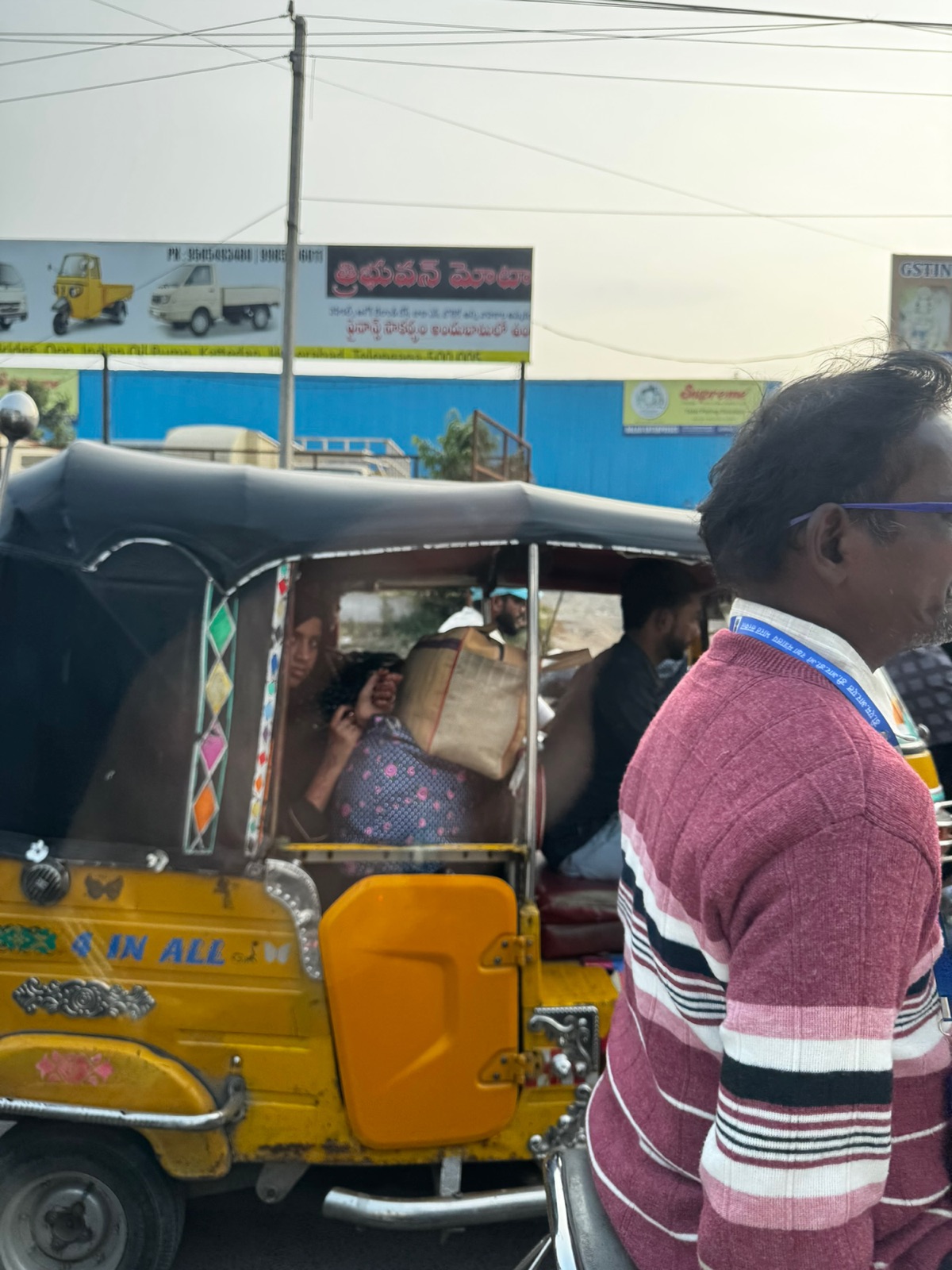Κίτρινο auto-rickshaw με την ένδειξη 4 IN ALL να μεταφέρει επιβάτες μπροστά από διαφημιστικές πινακίδες στα Τελούγκου