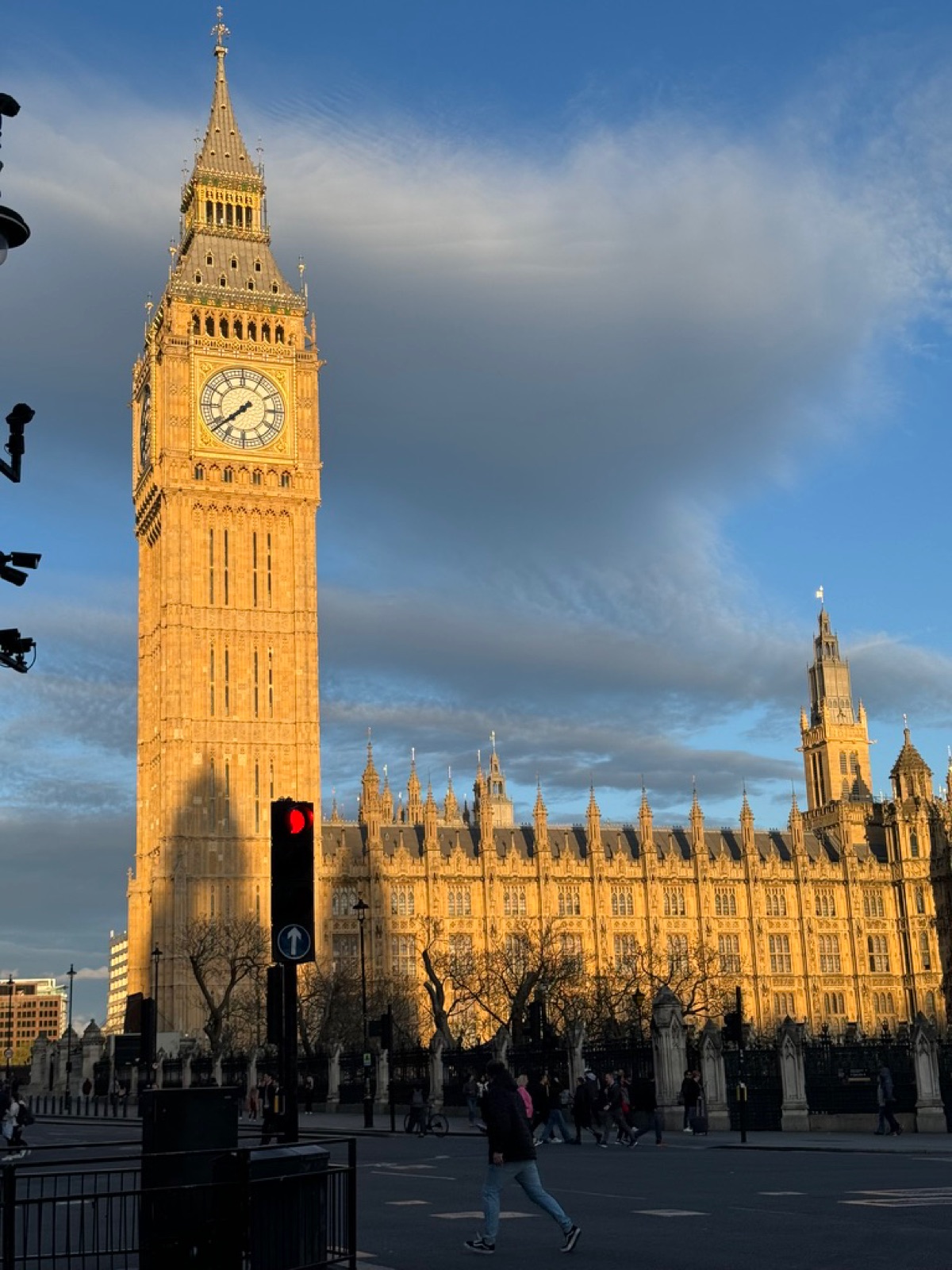 Το Big Ben και το κτίριο του Κοινοβουλίου να λάμπουν χρυσά στο βραδινό φως κάτω από γαλάζιο ουρανό, κόσμος να διασχίζει σε κόκκινο φανάρι πεζών