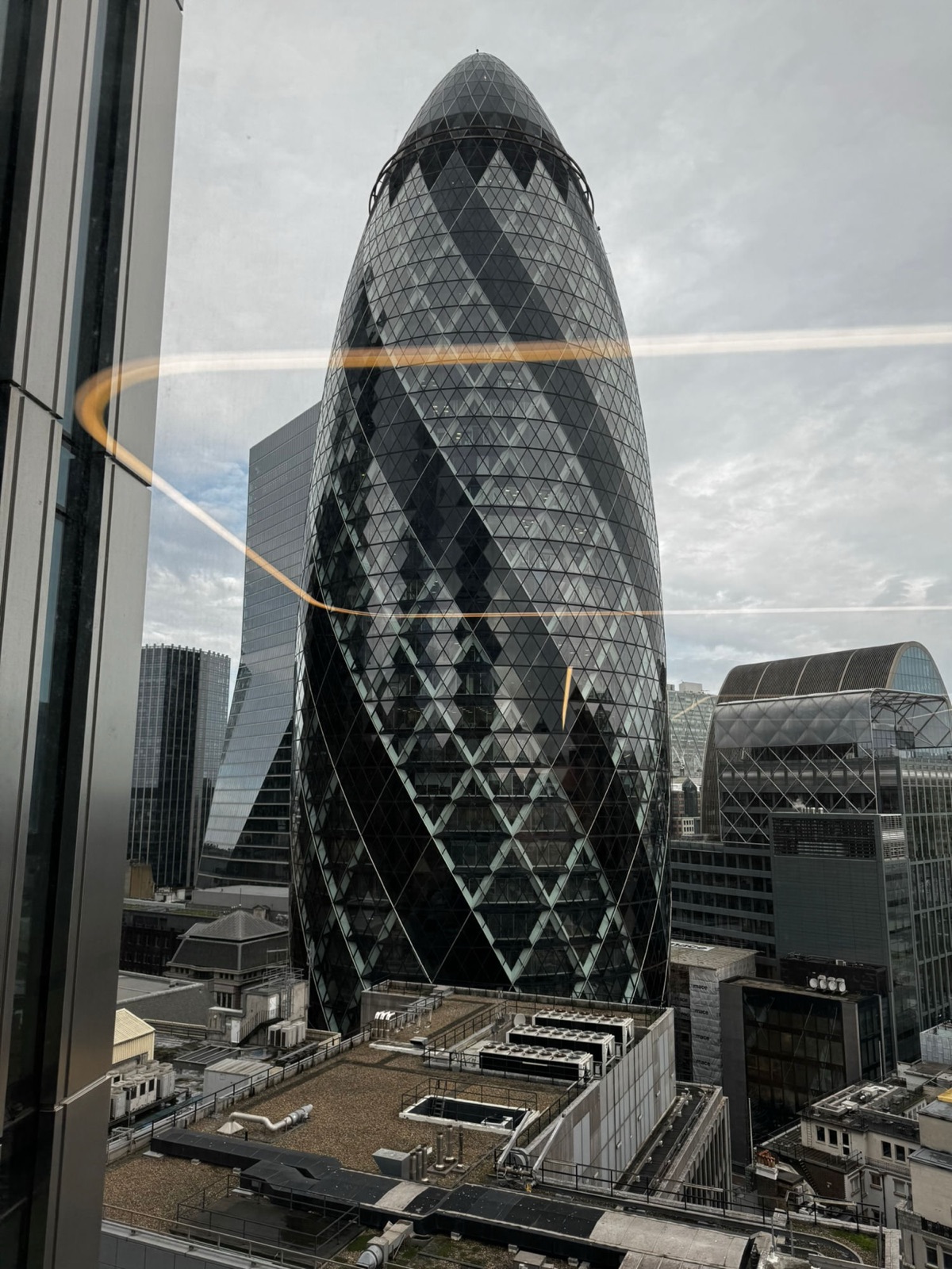 Το Gherkin ιδωμένο στο ίδιο ύψος από ένα κοντινό παράθυρο γραφείου, μια αντανάκλαση λάμπας να σχεδιάζει μια χρυσή έλλειψη στο γυαλί, μηχανοστάσια στέγης από κάτω