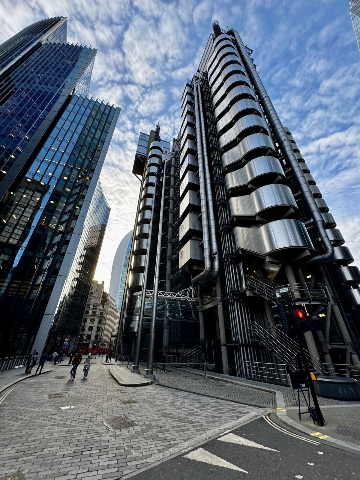 Το κτίριο Lloyd's από επίπεδο δρόμου κάτω από έναν λαμπερό γαλάζιο ουρανό με αραιά σύννεφα, ποδηλάτες να περνούν στον λιθόστρωτο δρόμο