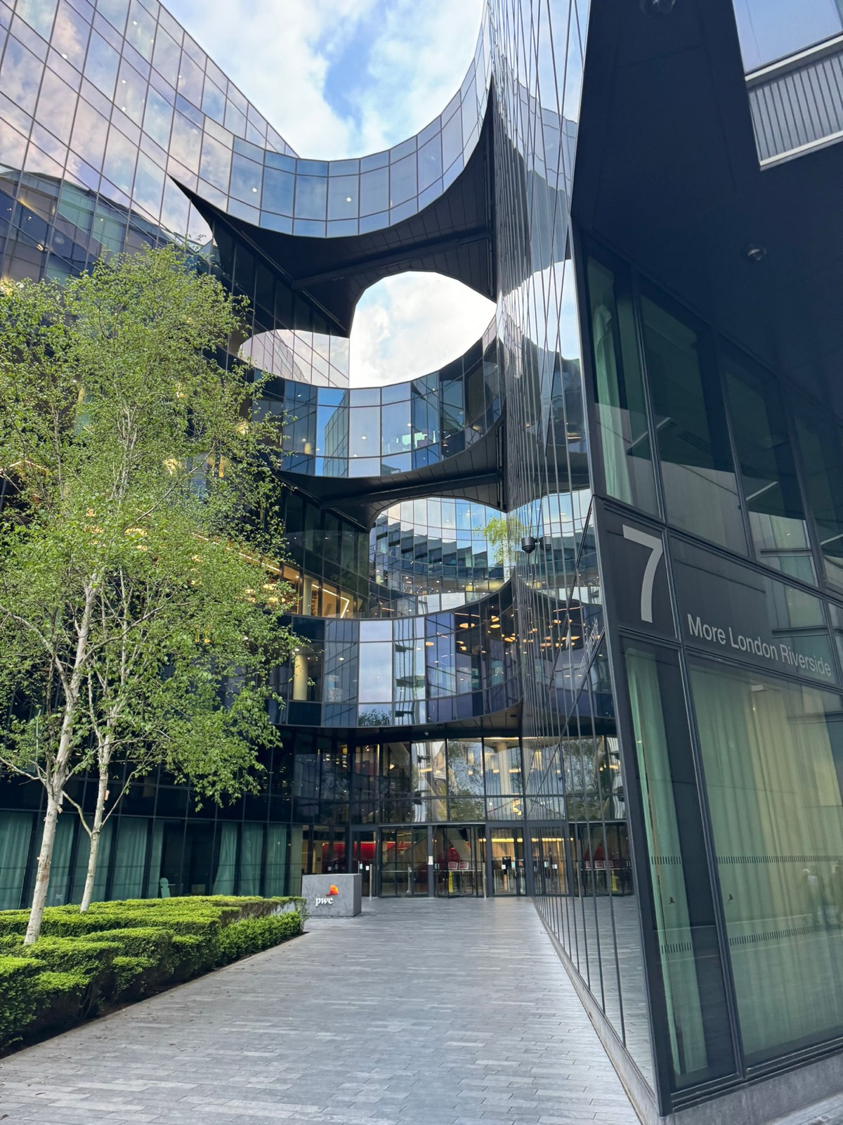 Το κτίριο PwC στο 7 More London Riverside, σκούρο γυαλί με καμπύλες εγκοπές που αποκαλύπτουν τους εσωτερικούς ορόφους, σημύδες μπροστά