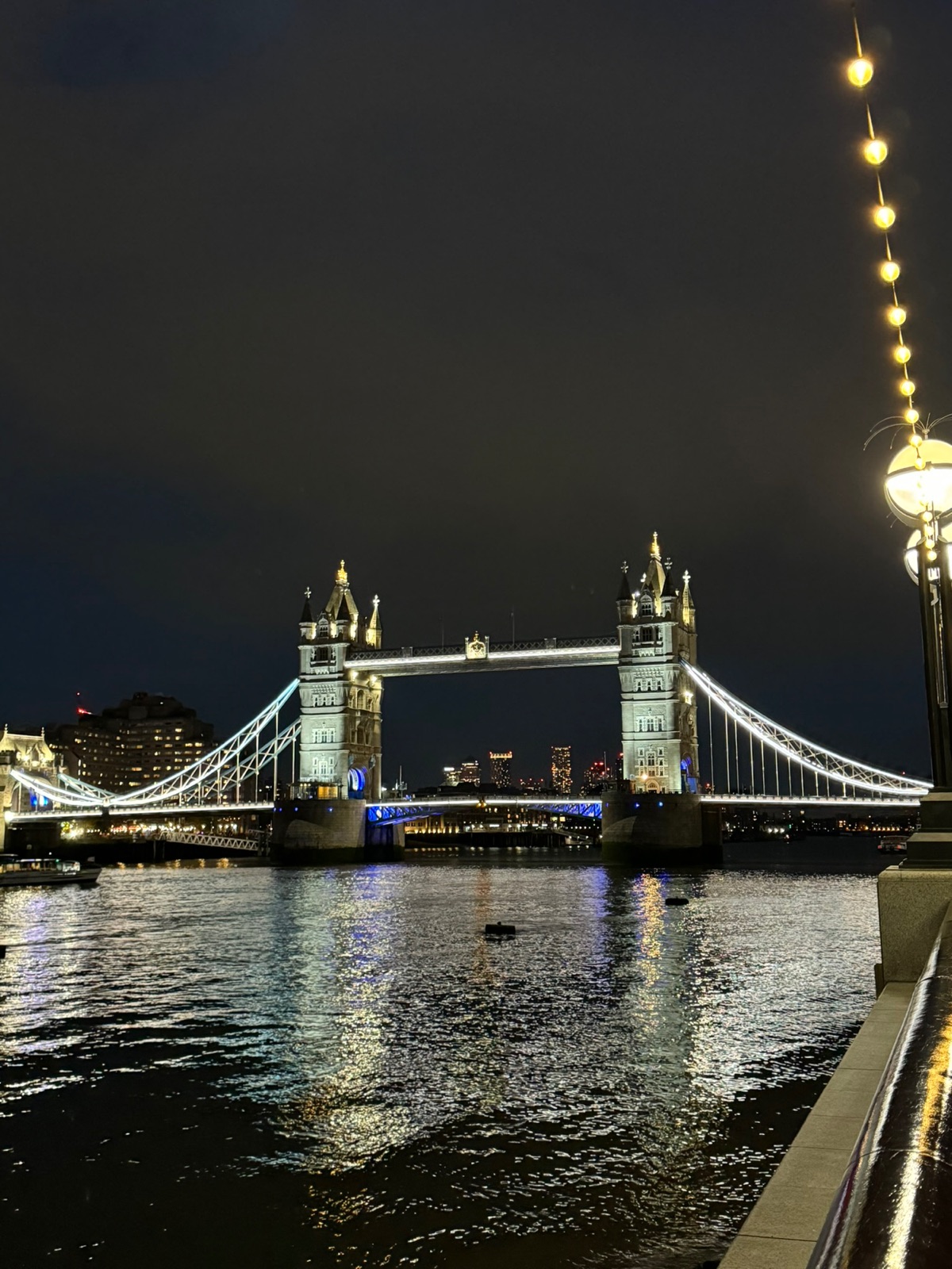 Η Tower Bridge πλήρως φωτισμένη τη νύχτα, οι αλυσίδες ανάρτησής της σχεδιασμένες σε λευκό φως, αντανακλάσεις σκορπισμένες στο σκοτεινό ποτάμι