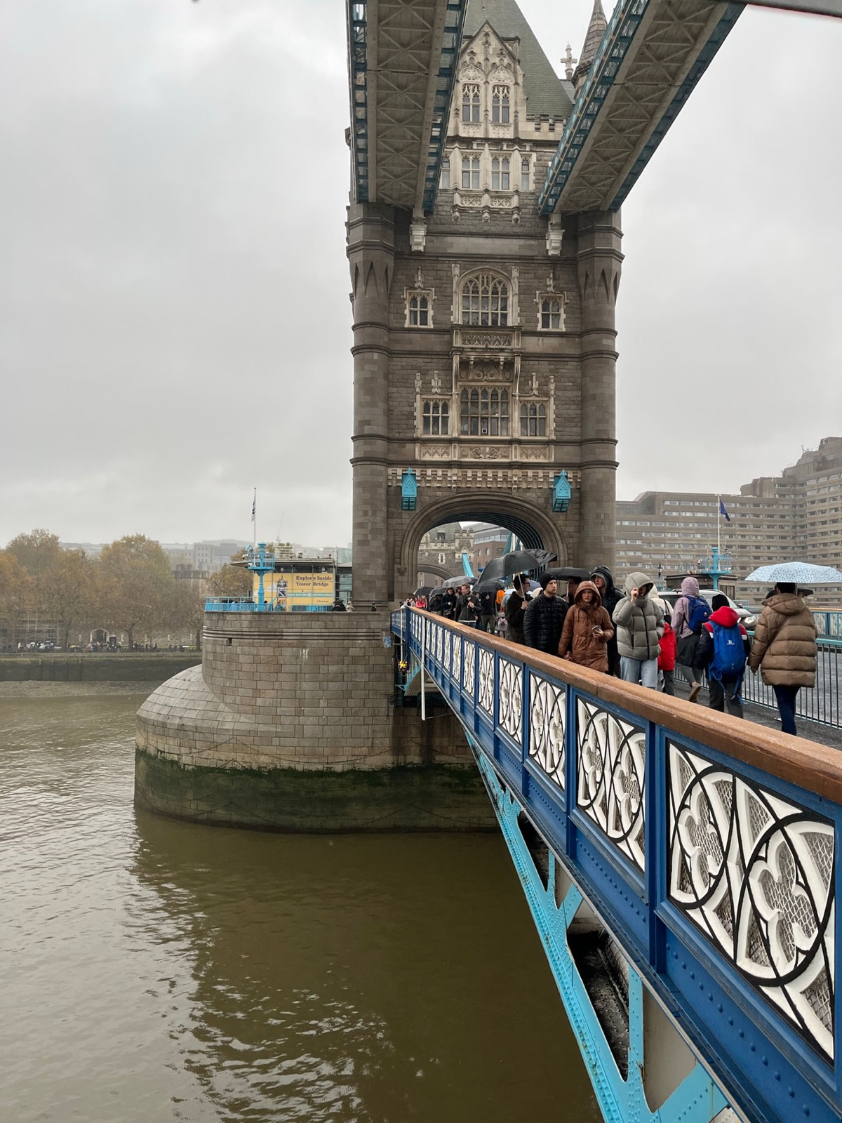 Τουρίστες με ομπρέλες να διασχίζουν την Tower Bridge μέσα σε δυνατή βροχή, ο πέτρινος πύργος και τα μπλε σιδερικά βρεγμένα και γκρίζα