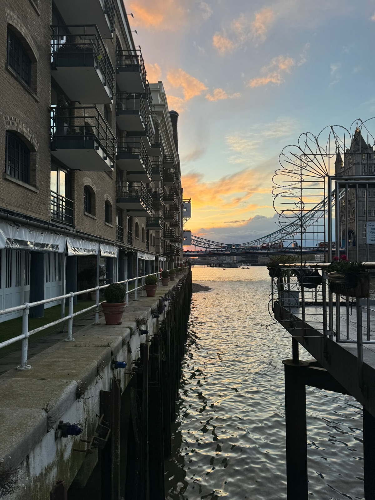Ηλιοβασίλεμα πίσω από την Tower Bridge ιδωμένο μέσα από ένα στενό κανάλι ανάμεσα σε κτίρια αποβάθρας, κόκκινα λεωφορεία να διασχίζουν τη γέφυρα, πορτοκαλιά σύννεφα να καθρεφτίζονται στο νερό