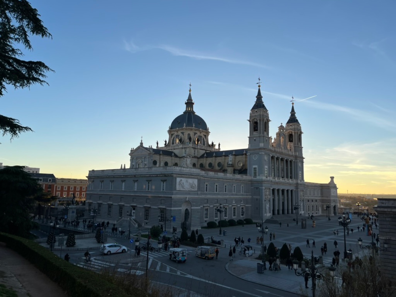 Ο καθεδρικός της Almudena σε πλατιά λήψη πάνω σε καθαρό απογευματινό ουρανό, ο γκρίζος τρούλος και τα δίδυμα καμπαναριά του πάνω από τη μακριά πρόσοψη προς το παλάτι, ένα ταξί και τουριστικά τουκ τουκ στη διάβαση από κάτω