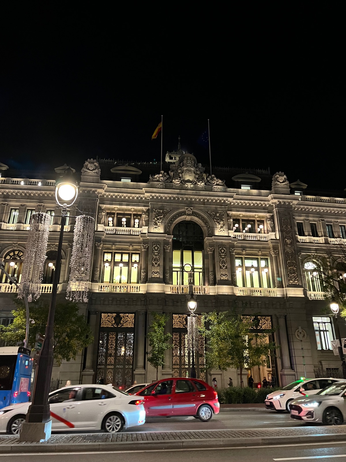 Η σκαλισμένη γκρίζα πέτρινη πρόσοψη της Banco de España φωτισμένη τη νύχτα, ένας στύλος τυλιγμένος με λαμπάκια και ταξί να περνούν μπροστά