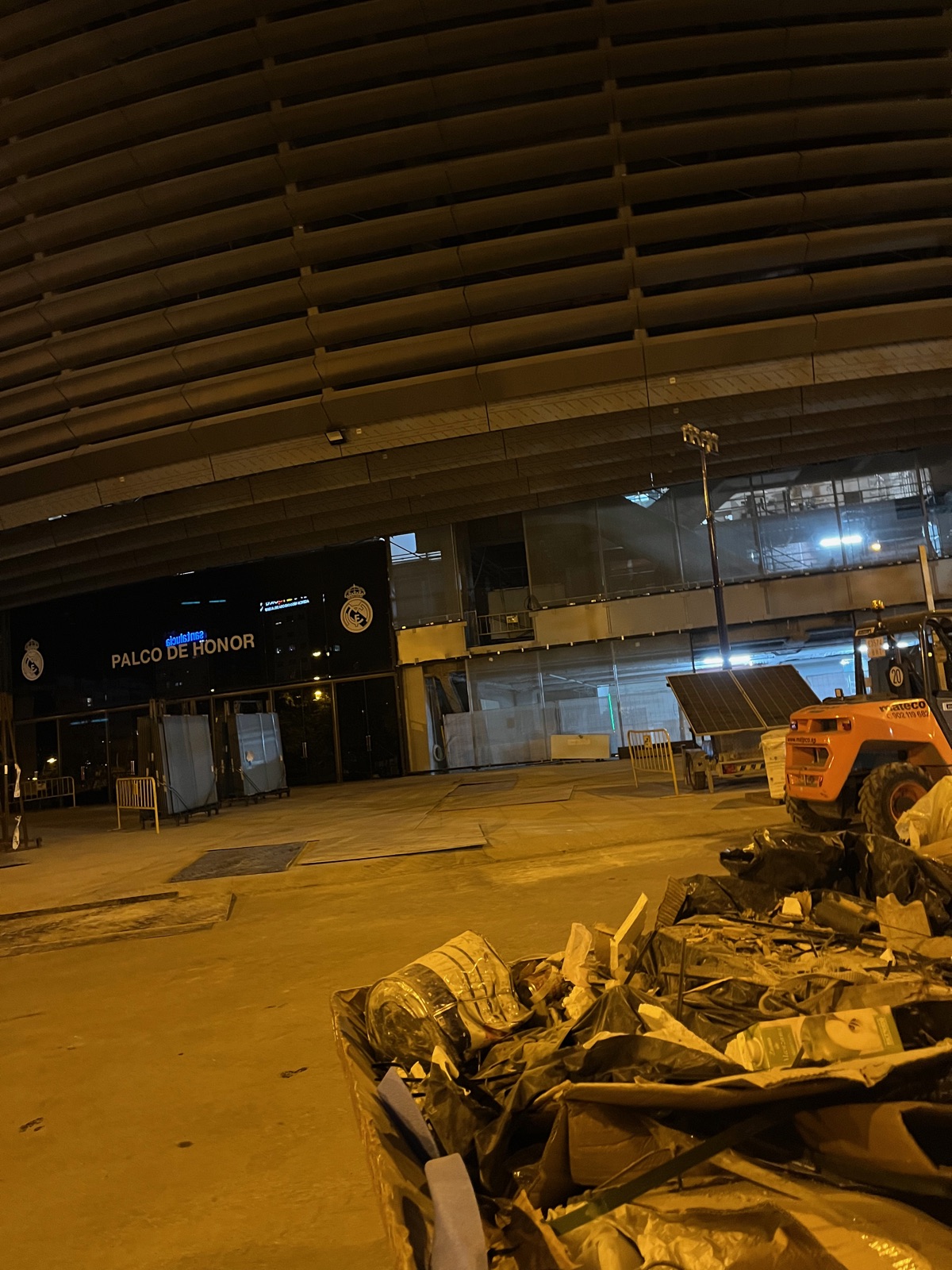 Μια φωτισμένη περιοχή εργοταξίου κάτω από την κατασκευή του σταδίου, μπάζα και ένας πορτοκαλί εκσκαφέας πίσω από περίφραξη