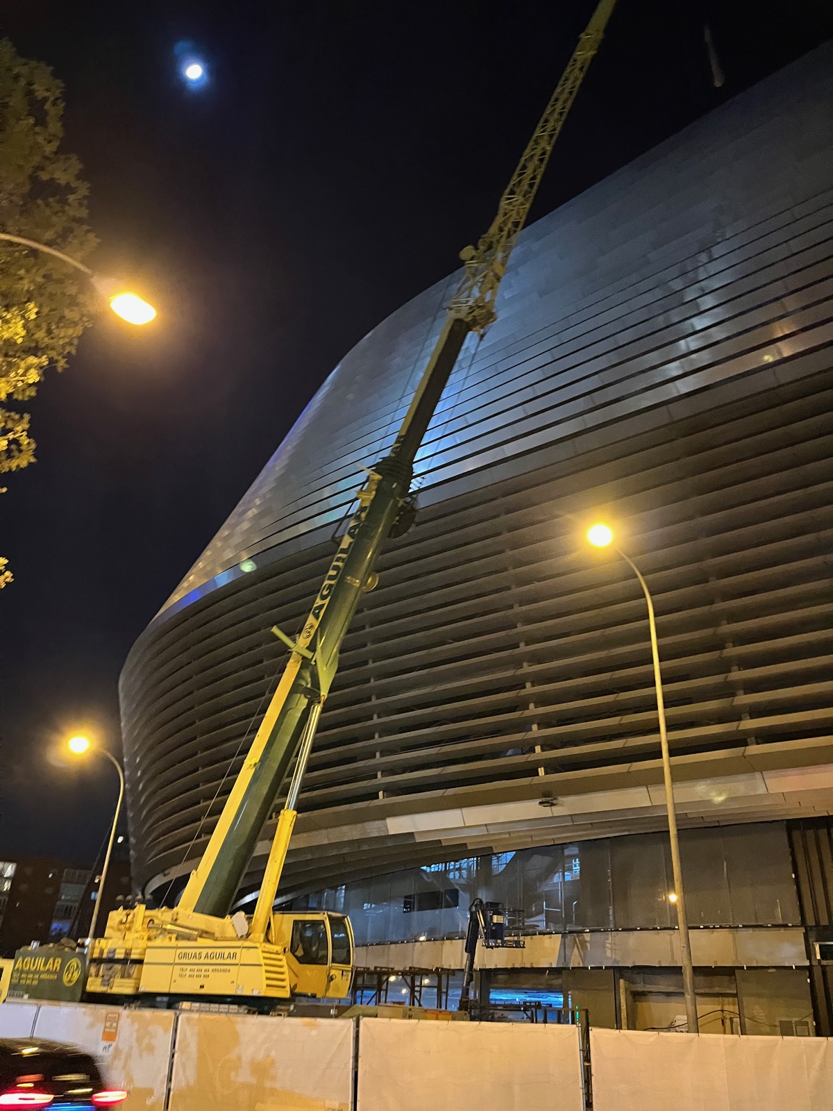 Ένας ψηλός κίτρινος γερανός να στέκεται πάνω από την καμπύλη ατσάλινη πρόσοψη του Μπερναμπέου τη νύχτα, το φεγγάρι μικρό στον ουρανό