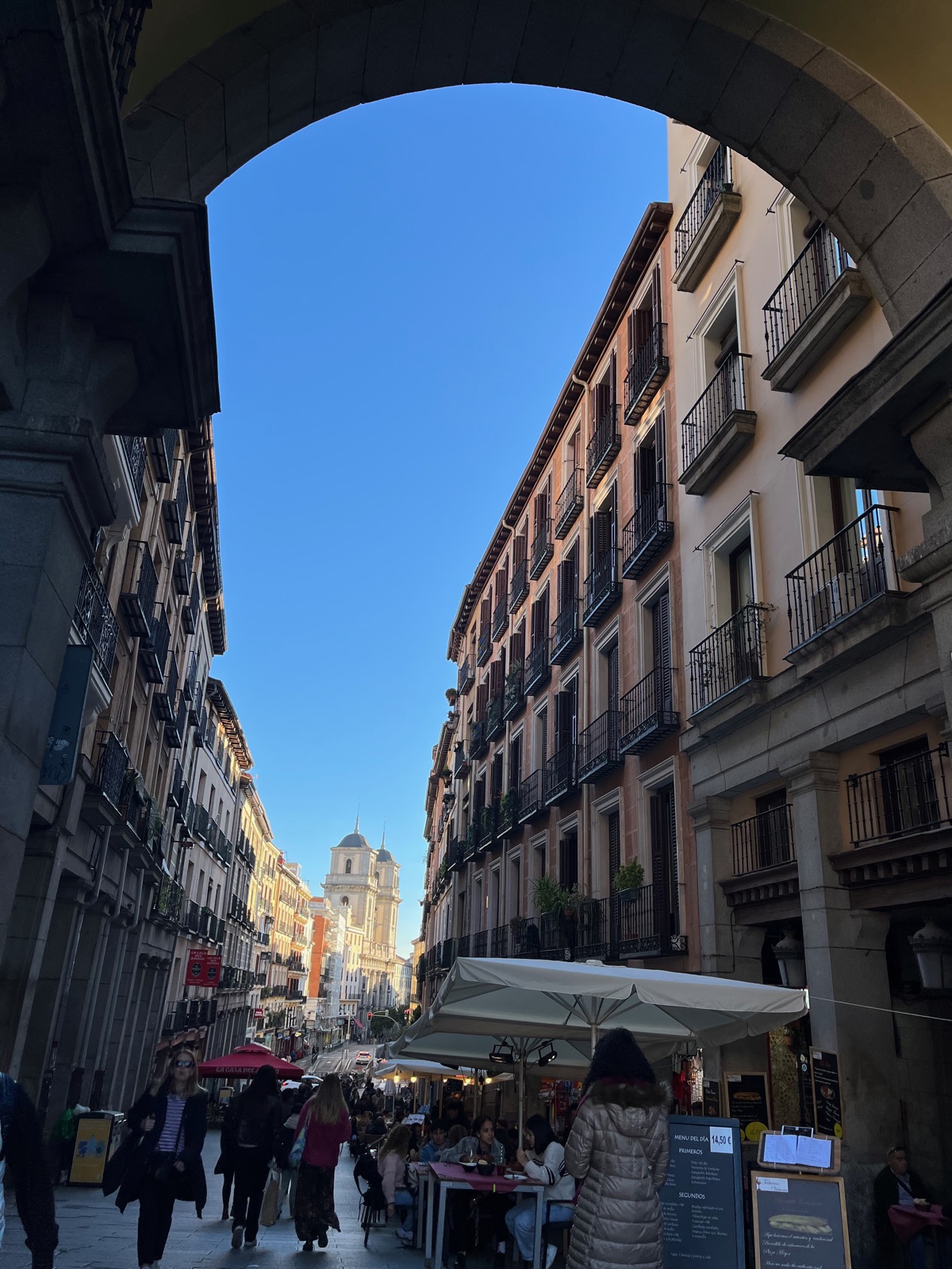 Θέα μέσα από μια πέτρινη καμάρα της Plaza Mayor προς την Calle de Toledo, βεράντες καφέ και πεζοί στον δρόμο και ένας τρούλος εκκλησίας στο βάθος