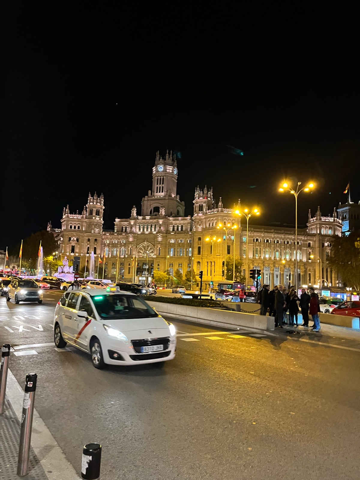 Το Palacio de Cibeles φωτισμένο τη νύχτα από την απέναντι πλευρά της πλατείας, ένα λευκό ταξί της Μαδρίτης με την κόκκινη διαγώνια λωρίδα του να περνάει στο πρώτο πλάνο και κίνηση σταματημένη δίπλα στο σιντριβάνι