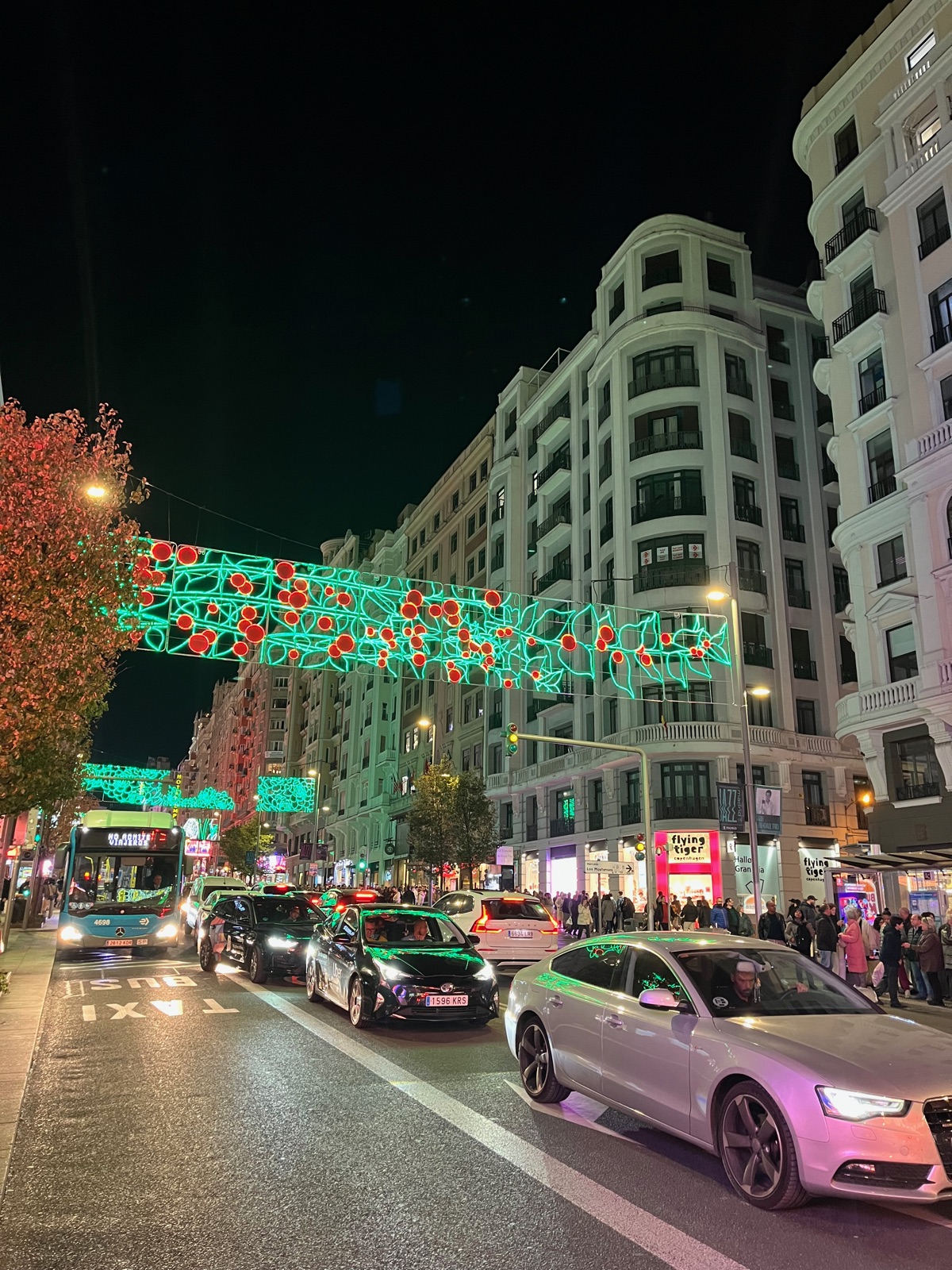 Πράσινες χριστουγεννιάτικες γιρλάντες με κόκκινες τελείες τεντωμένες πάνω από τη Gran Vía τη νύχτα, αυτοκίνητα και ένα λεωφορείο στον δρόμο που μοιάζει βρεγμένος ανάμεσα σε λευκά πέτρινα κτίρια