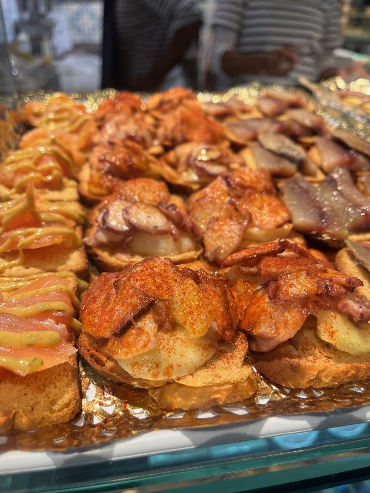 Ένας δίσκος με pintxos πάνω σε φέτες ψωμιού με ψητό κρέας και σάλτσα κάτω από ζεστό φως πάγκου σε στάντ αγοράς