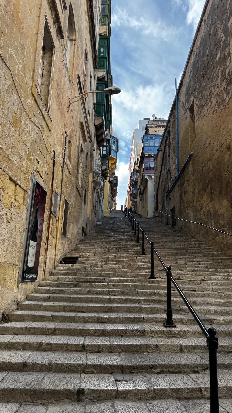 Ένας μακρύς άδειος σκαλωτός δρόμος ανάμεσα σε ασβεστολιθικά κτίρια, μαύρα σιδερένια κάγκελα στη μέση, πράσινα και μπλε βαμμένα ξύλινα μπαλκόνια να προεξέχουν από τις προσόψεις ψηλά