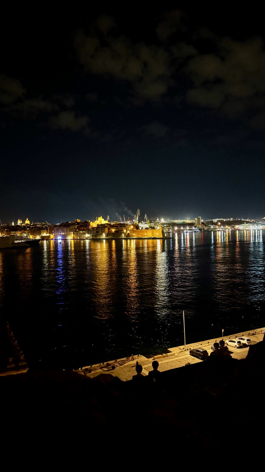 Το Μεγάλο Λιμάνι τη νύχτα από ψηλά, οι φωτισμένες οχυρώσεις και τα κτίρια της απέναντι ακτής καθρεφτισμένα σε μακριές λωρίδες στο μαύρο νερό, γερανοί ναυπηγείου στο βάθος