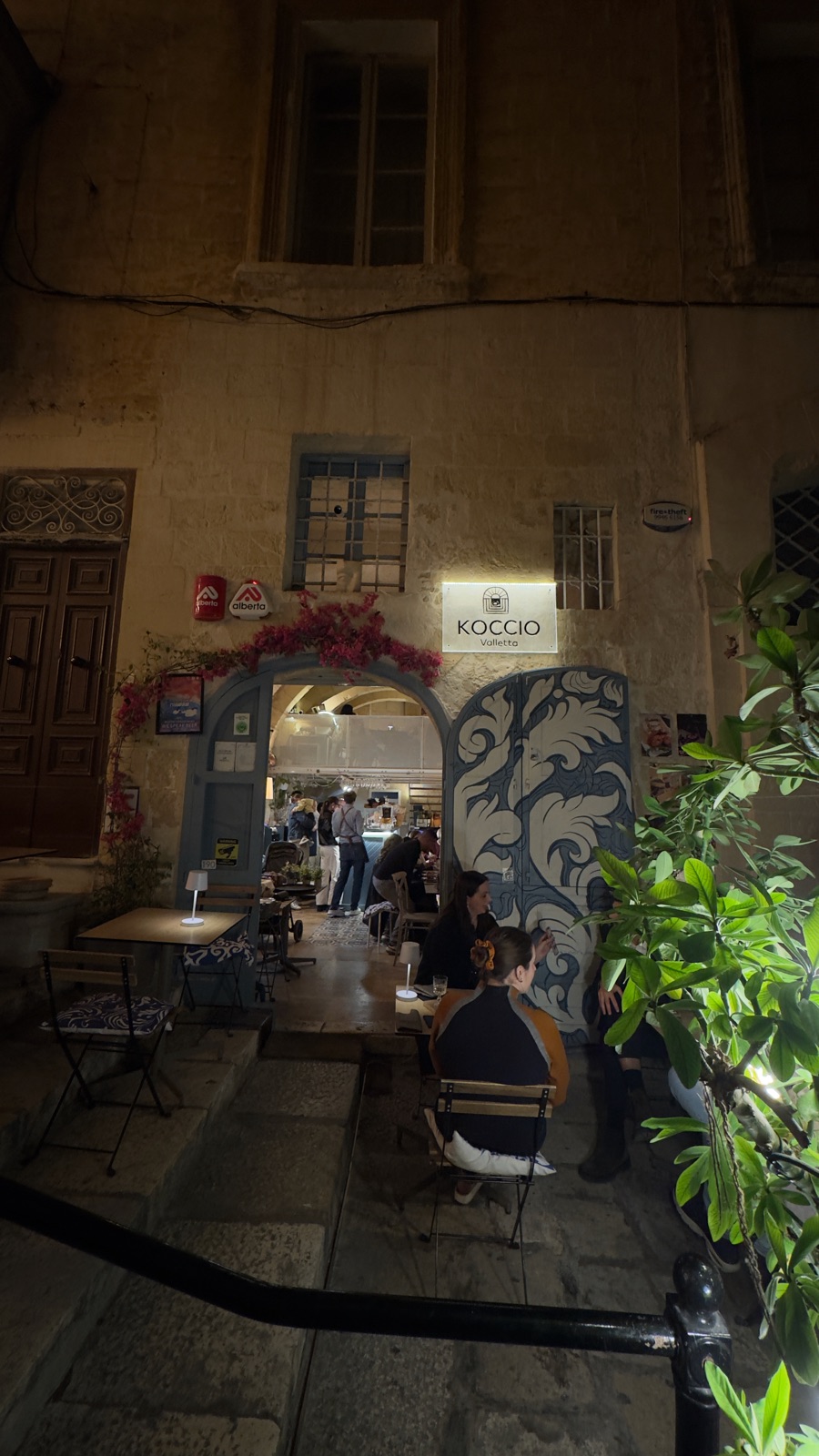 Ένα καφέ σε αυλή τη νύχτα πίσω από μια τοξωτή είσοδο με μπλε και λευκές ζωγραφισμένες πόρτες, μια φωτισμένη πινακίδα που γράφει KOCCIO Valletta και βουκαμβίλια πάνω από την καμάρα, κόσμος σε μικρά τραπέζια έξω