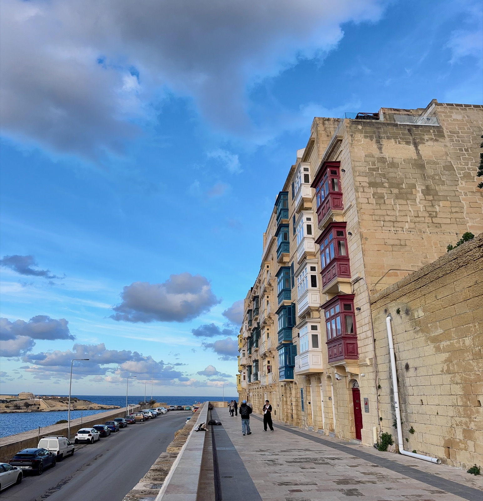 Μια παραλιακή σειρά ασβεστολιθικών σπιτιών με στοιβαγμένα ξύλινα μπαλκόνια σε κόκκινο, λευκό και μπλε, ένας πλατύς πλακόστρωτος περίπατος μπροστά και η ανοιχτή θάλασσα πέρα από τον δρόμο