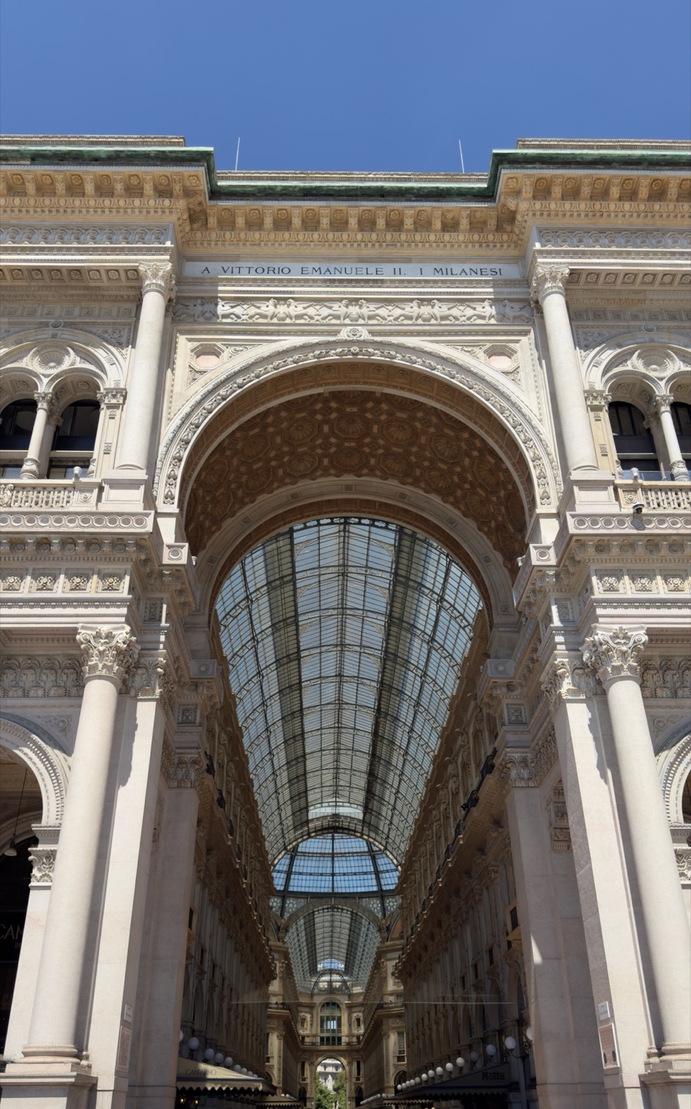 Η αψίδα εισόδου της Galleria Vittorio Emanuele II από την Piazza del Duomo, η αφιέρωση στον Vittorio Emanuele II σκαλισμένη στο ανώτερο τμήμα και ο γυάλινος θόλος να φαίνεται μέσα από την αψίδα