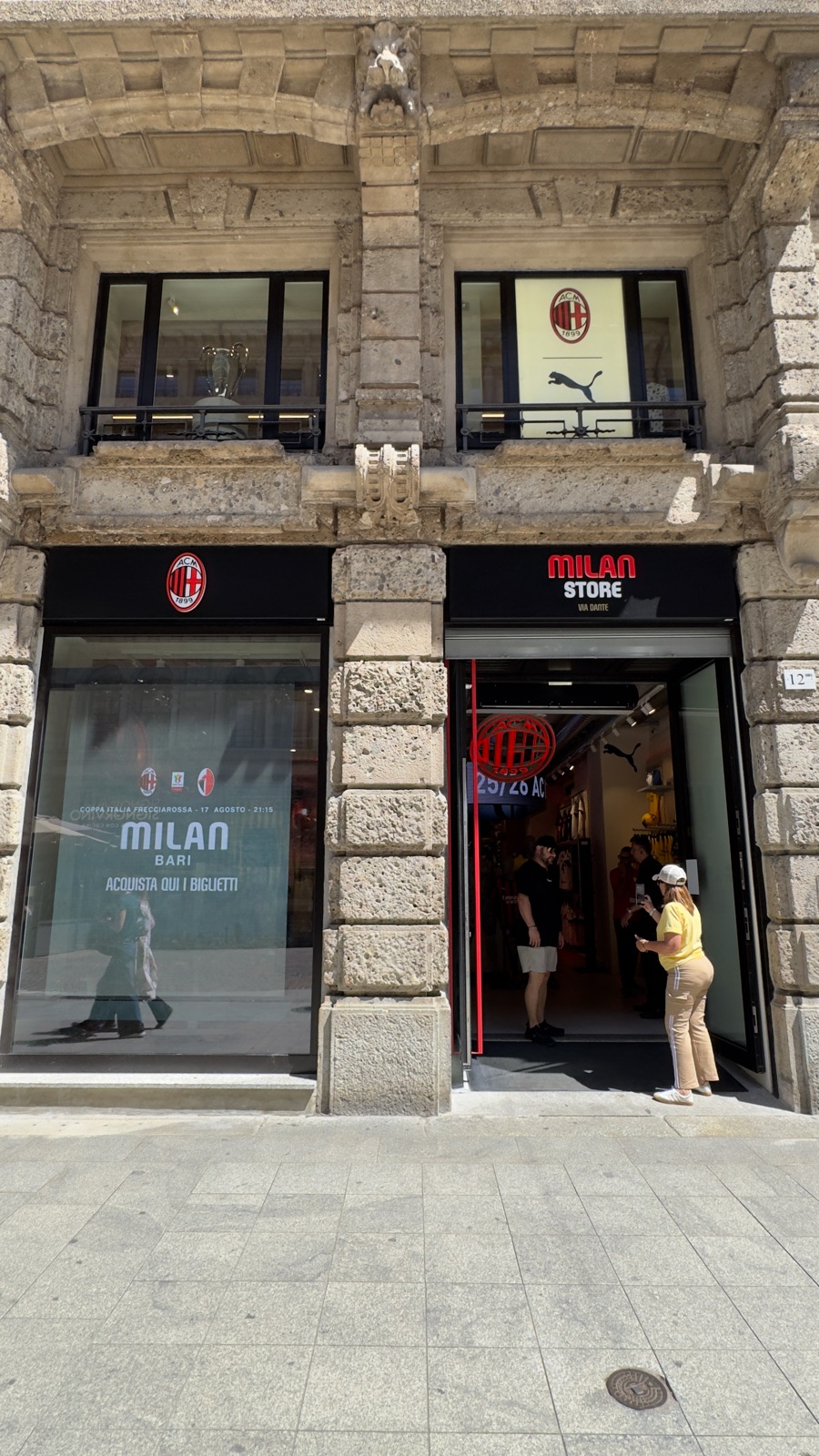 Η πρόσοψη του AC Milan Store λίγες πόρτες πιο κάτω στη Via Dante, σήματα του συλλόγου πάνω από την είσοδο, ένα τρόπαιο στο παράθυρο του πρώτου ορόφου και μια αφίσα εισιτηρίων στη βιτρίνα