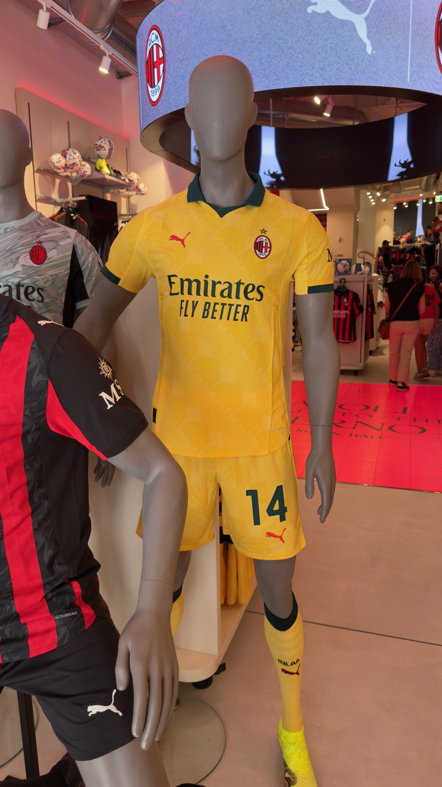 Μια κούκλα με κίτρινη εμφάνιση της Puma και σκούρα πράσινα τελειώματα μέσα στο κατάστημα της Μίλαν, το σήμα του συλλόγου σε κύκλο οθονών από πάνω και ράφια με μπάλες πίσω