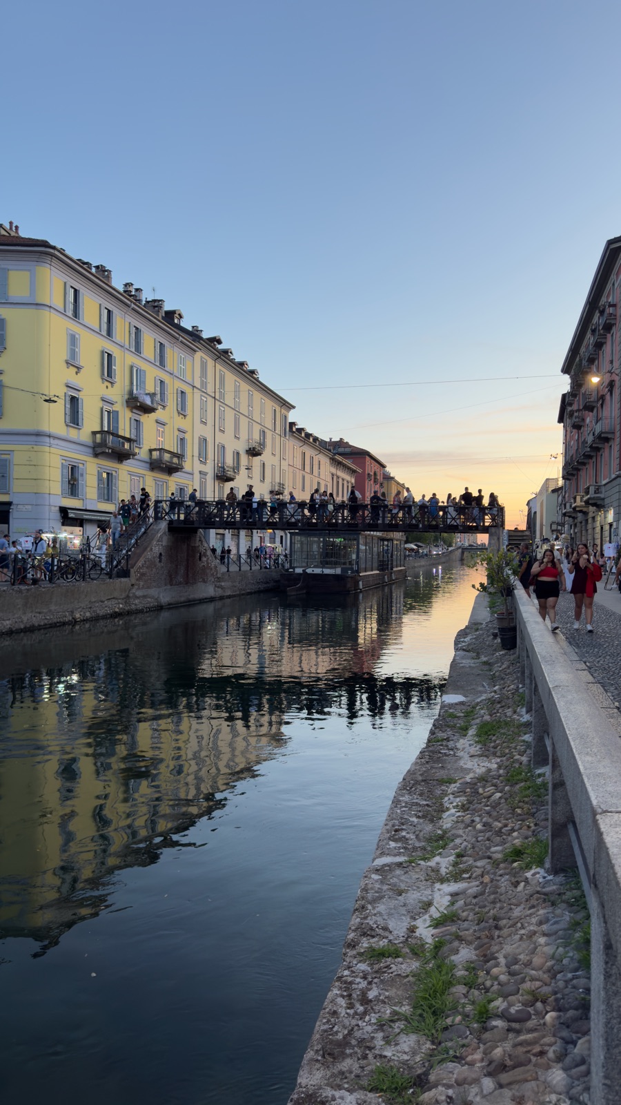 Μια σιδερένια πεζογέφυρα πάνω από το Naviglio Grande γεμάτη κόσμο σε σιλουέτα το σούρουπο, μια μαούνα με γυάλινη οροφή δεμένη από κάτω και ο ουρανός να λάμπει πορτοκαλής πίσω από τα κτίρια
