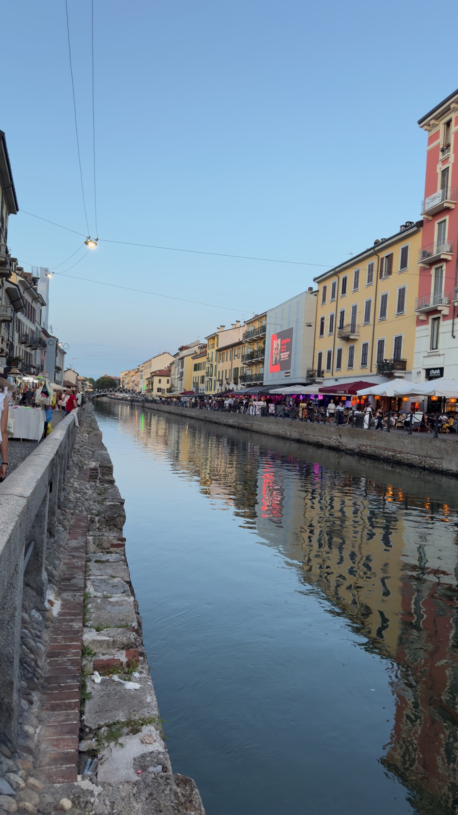 Το Naviglio Grande τη γαλάζια ώρα από την πέτρινη άκρη του μονοπατιού, η απέναντι όχθη φωτισμένη από μπαρ και μια κόκκινη διαφημιστική πινακίδα, τα φώτα διπλασιασμένα στο σκοτεινό νερό