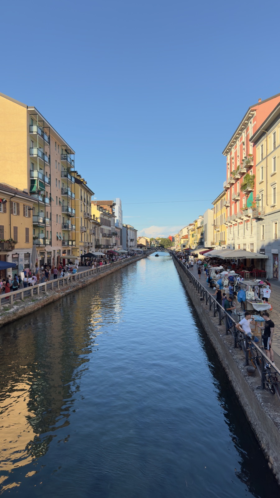 Το Naviglio Grande σε χρυσό βραδινό φως, ένα μακρύ ίσιο κανάλι ανάμεσα σε κίτρινα και κεραμιδί κτίρια, πάγκοι αγοράς και ομπρέλες οστερίας στη δεξιά όχθη και κόσμος και στα δύο μονοπάτια