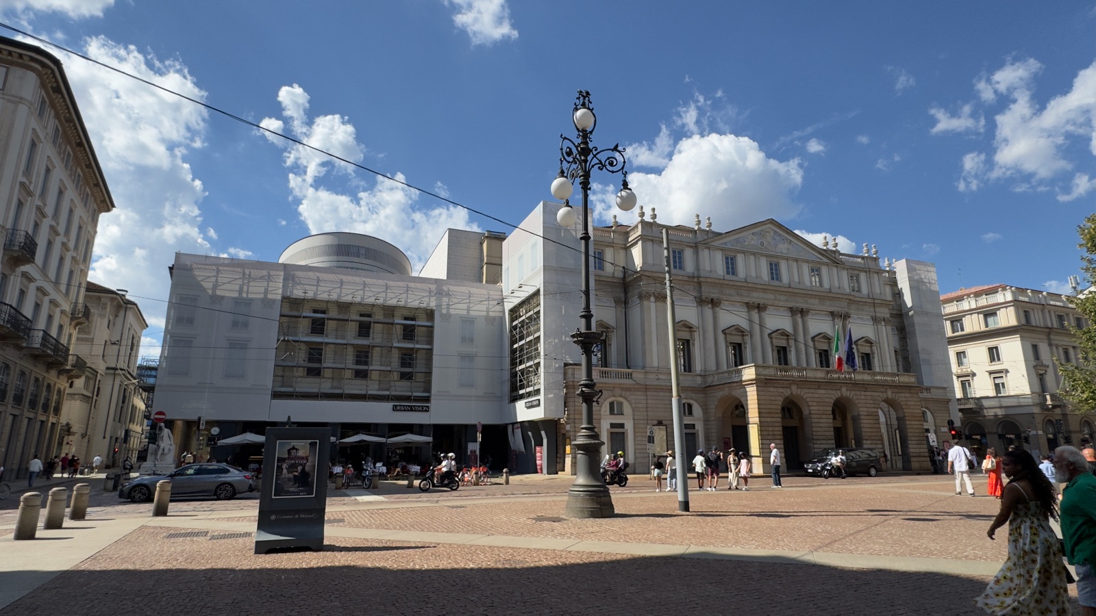 Η Piazza della Scala κάτω από γαλάζιο ουρανό με σκόρπια σύννεφα, η πρόσοψη του Teatro alla Scala με ιταλικές και ευρωπαϊκές σημαίες, το μισό τετράγωνο τυλιγμένο σε τυπωμένο ύφασμα ανακαίνισης, ένα διακοσμημένο φανάρι στο πρώτο πλάνο