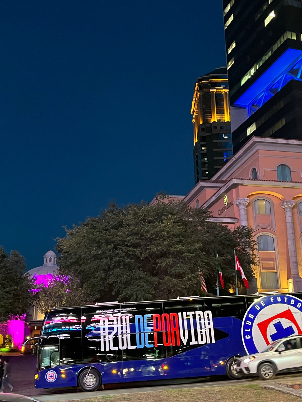 Το μπλε πούλμαν της ομάδας Cruz Azul με τα γράμματα Azul De Por Vida παρκαρισμένο έξω από ένα ξενοδοχείο τη νύχτα, ένας φωτισμένος πύργος πίσω