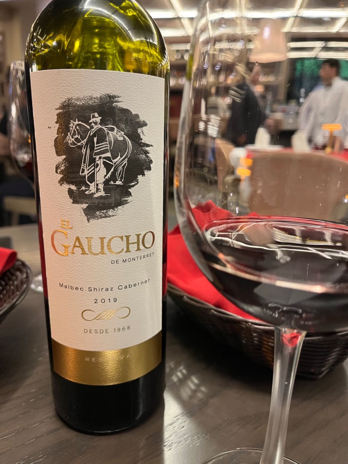 Ένα μπουκάλι El Gaucho de Monterrey Malbec Shiraz Cabernet 2019 δίπλα σε ένα σερβιρισμένο ποτήρι κόκκινο κρασί στο τραπέζι