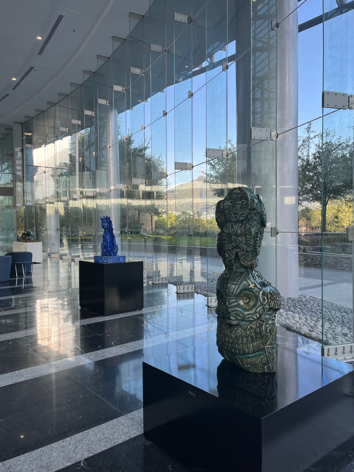 Ένα λόμπι με γυάλινο τοίχωμα, με μια κοβαλτένια κεραμική φιγούρα και ένα πράσινο σκαλισμένο τοτέμ σε μαύρα βάθρα, κήποι ορατοί έξω