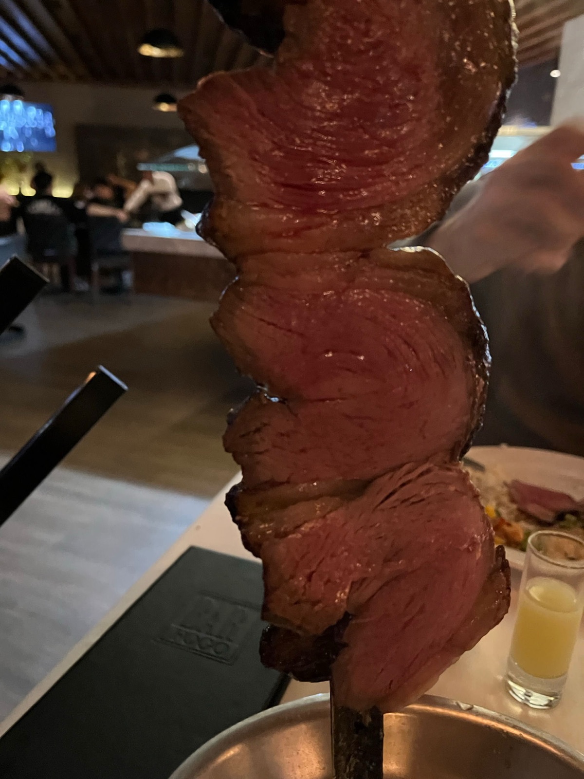 Ένα σουβλί με ροδαλές φέτες picanha κρατημένο πάνω από το τραπέζι στην churrascaria, το ανάγλυφο μενού Bar Fogo από κάτω