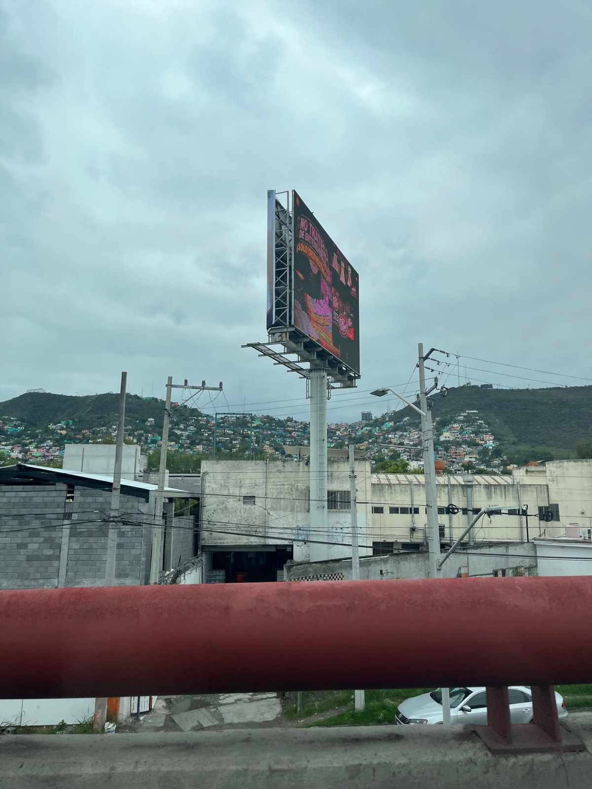 Μια colonia σε πλαγιά με πολύχρωμα σπίτια, ιδωμένη πάνω από μια κόκκινη μπάρα αυτοκινητόδρομου, κάτω από πυκνά σύννεφα