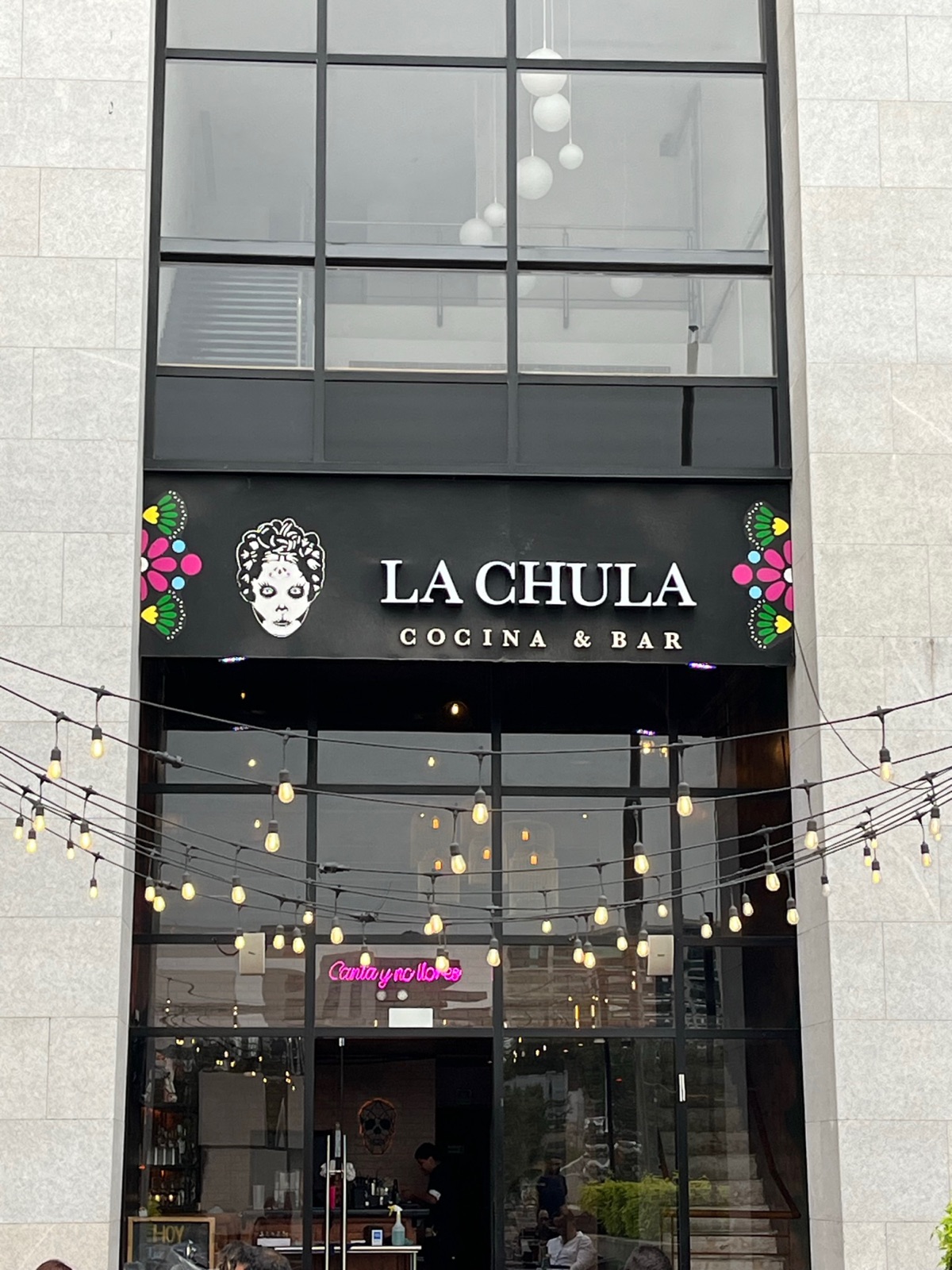 Η πρόσοψη του La Chula Cocina and Bar με ένα λογότυπο νεκροκεφαλής ζάχαρης, γιρλάντες φώτων και μια ροζ νέον πινακίδα μέσα