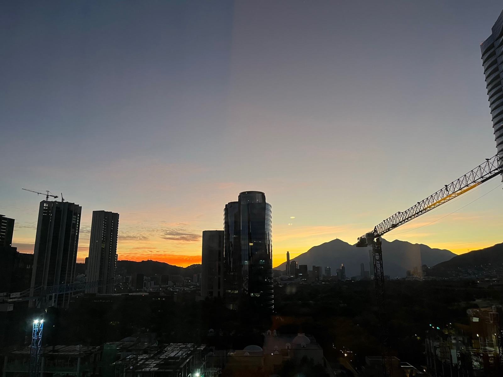 Ηλιοβασίλεμα πάνω από το Valle Oriente από ψηλό παράθυρο, πυργογερανοί και γυάλινοι ουρανοξύστες σε σιλουέτα πάνω σε πορτοκαλί ουρανό και τα βουνά