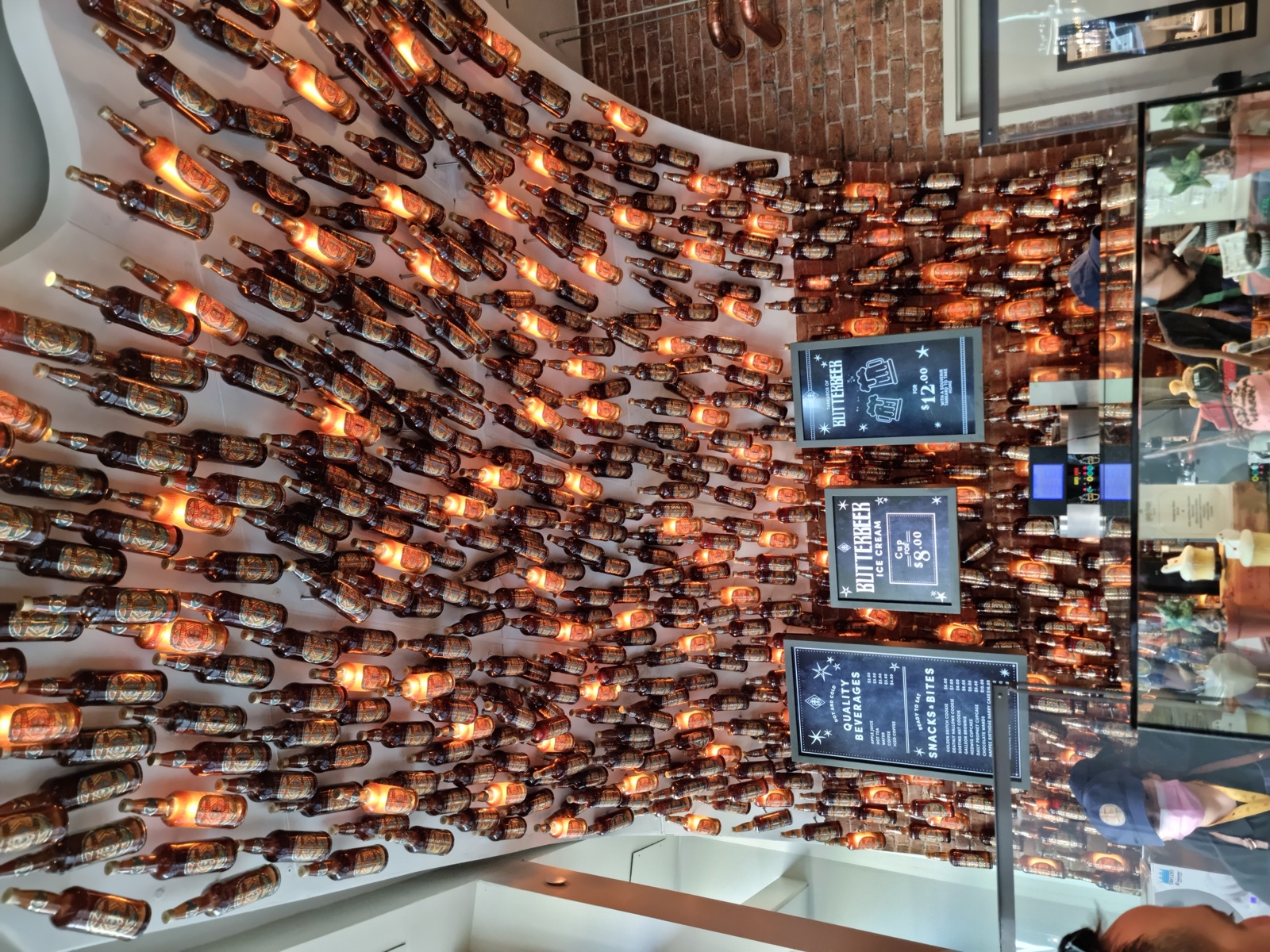 A vaulted ceiling hung with hundreds of illuminated amber glass bottles above the counter of the Butterbeer Bar in Harry Potter New York, with menu boards reading BUTTERBEER