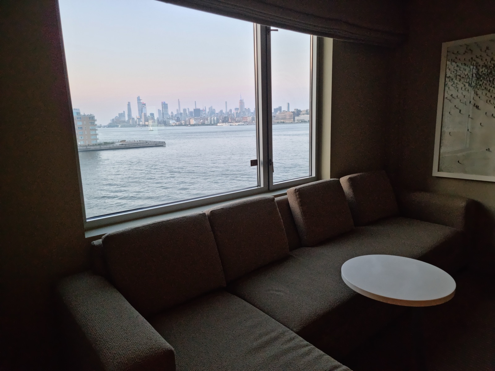 A hotel room sofa and low table in front of a wide window looking out over the Hudson to the Manhattan skyline at dusk