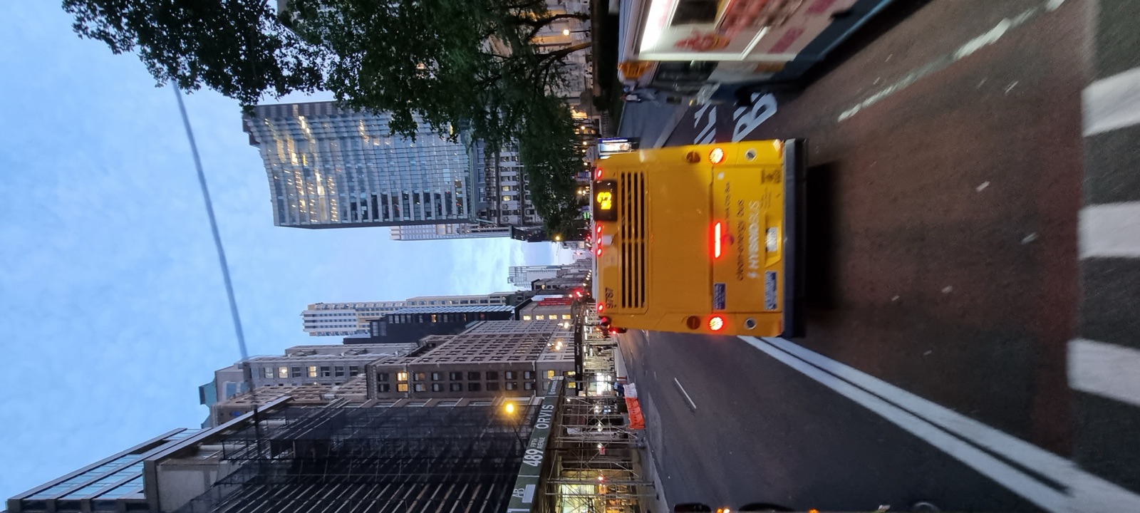 A yellow school bus driving away up a wide Manhattan avenue at dusk with lit buildings on both sides